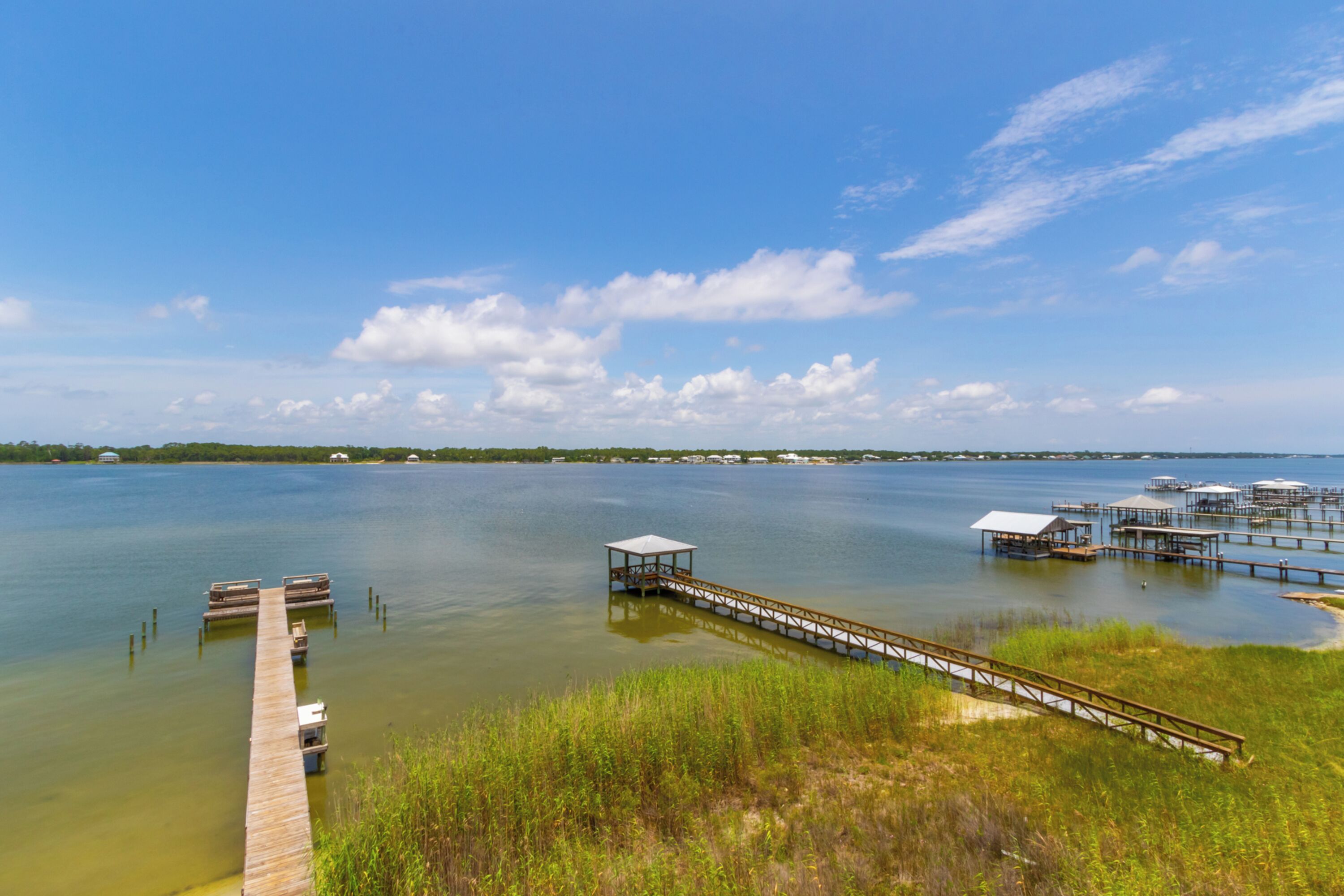 A waterfront property with a wooden dock extending into calm water, surrounded by grassy areas and distant buildings.