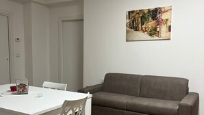 A modern living room with a white dining table, two white chairs, a brown sofa, and a wall-mounted air conditioner. - Savoy Apartment (Bari)