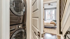 A laundry room with stacked washers and dryers, a hallway leading to a bedroom with a bed and a window.
