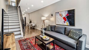 A modern living room with a grey sofa, a coffee table, a red patterned rug, a staircase with black railings, and a large abstract painting on the wall.