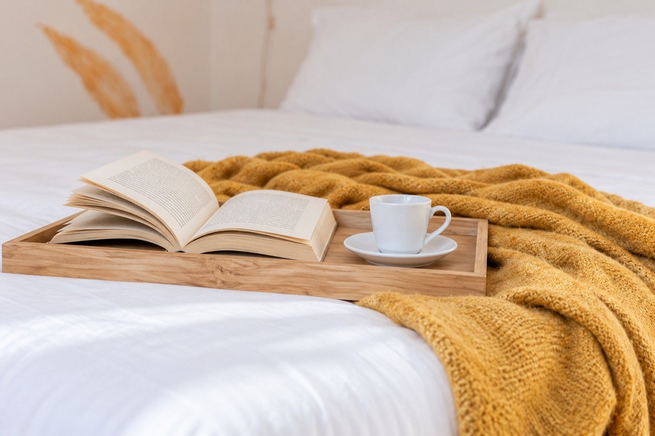 A bed with a white comforter, a mustard-colored knitted blanket, an open book, and a white cup on a wooden tray.