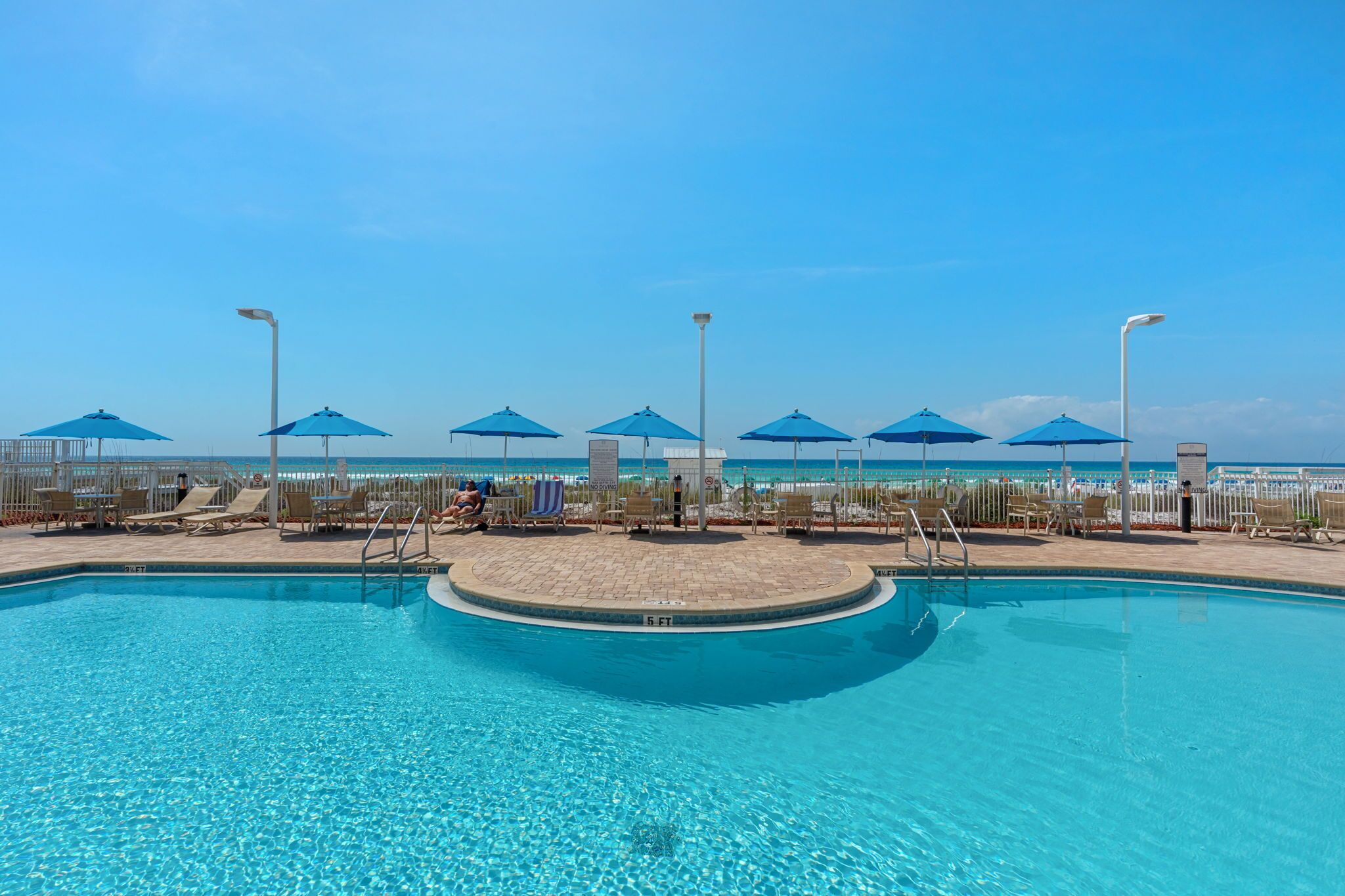 Piscina com espreguiçadeiras e guarda-sóis, céu claro e vista para a praia.
