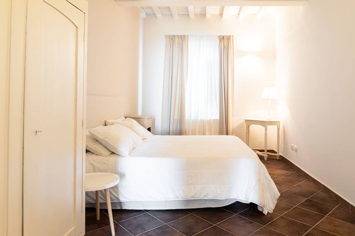 Un dormitorio con cama, mesita de noche, lámpara y ventana con cortinas.