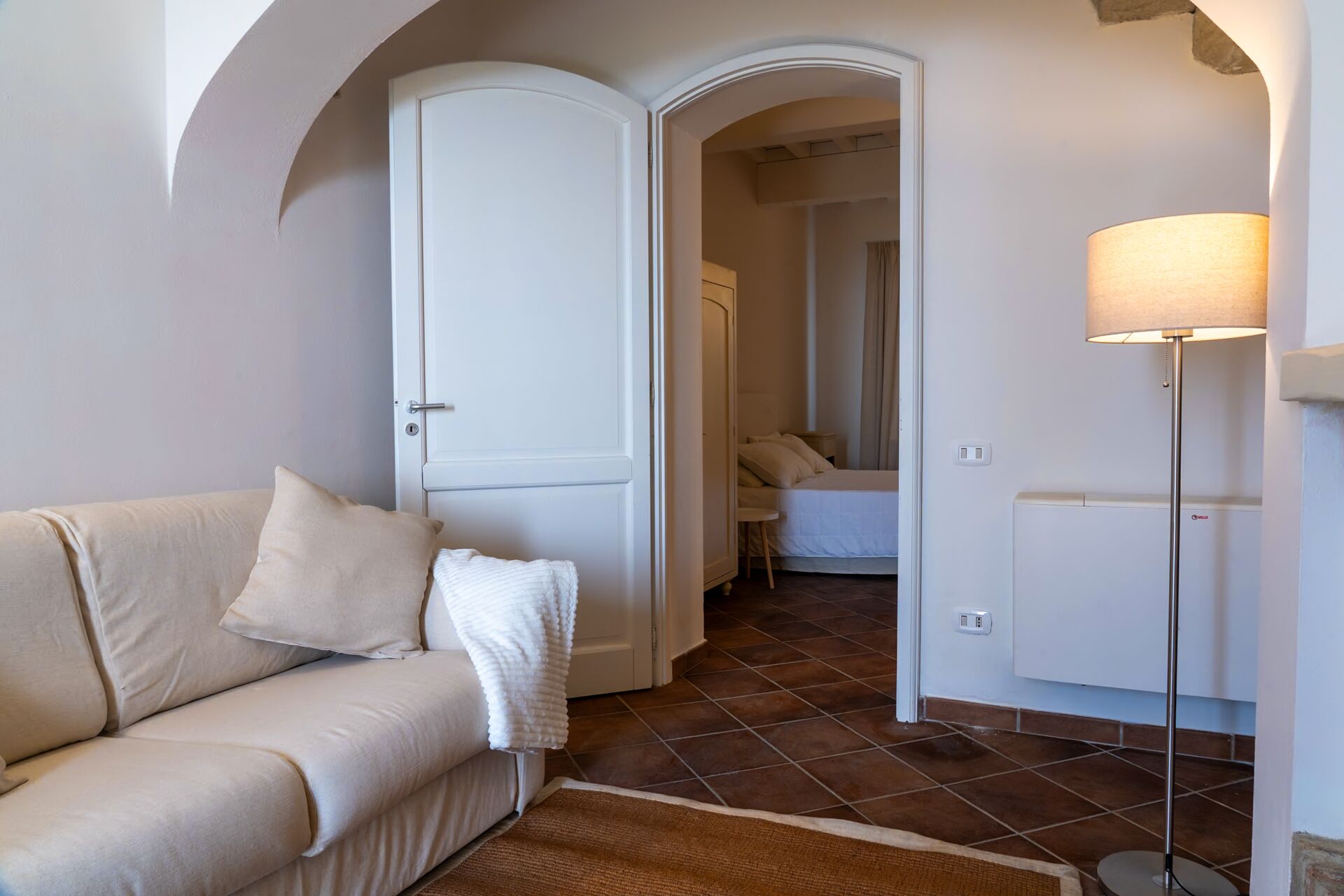 Een kamer met een beige bank, een witte deur, een bed met witte lakens, een vloerlamp en een radiator.