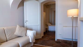 Een kamer met een beige bank, een witte deur, een bed met witte lakens, een vloerlamp en een radiator.