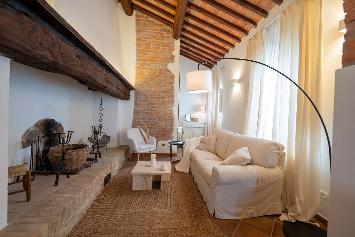 Una sala de estar acogedora con un sofá blanco, una mesa de centro y una chimenea. Se aprecia una pared de ladrillo y vigas de madera en el techo.