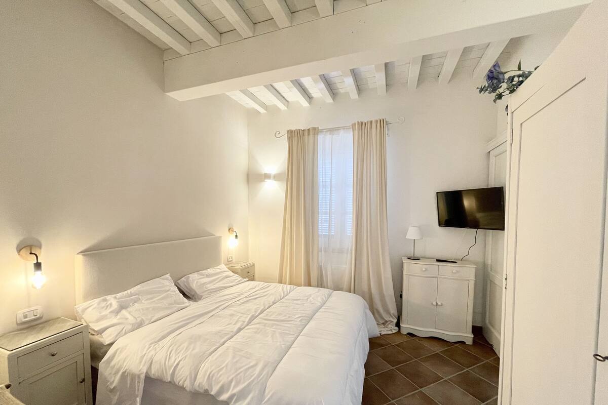 Un dormitorio con cama, mesitas de noche, televisión y ventana con cortinas.