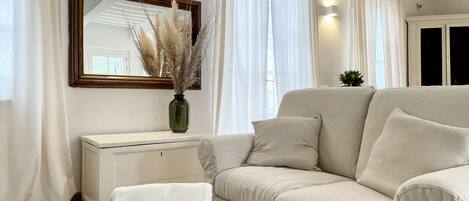 A modern living room with a beige sofa, a white ottoman, a green vase with pampas grass, and a large mirror on the wall.