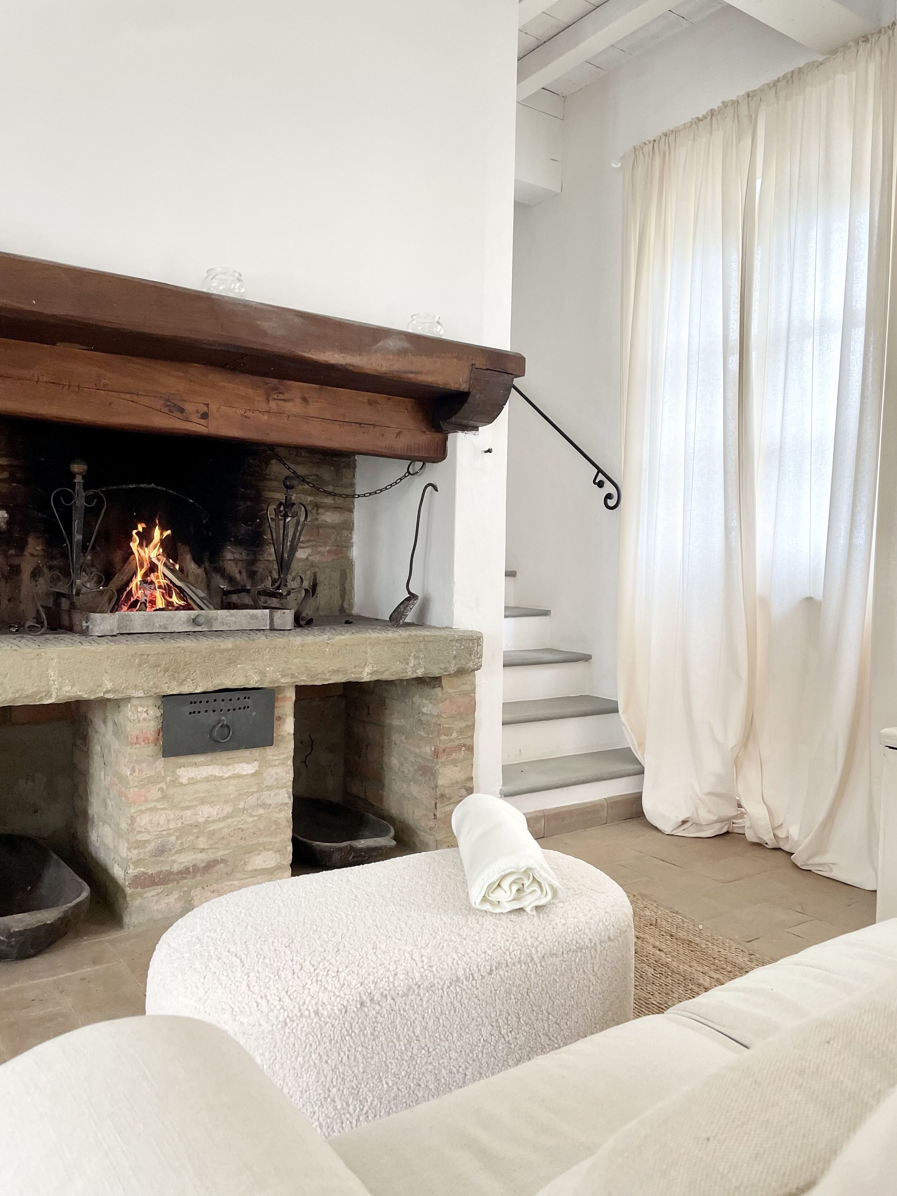 Un salon chaleureux avec une cheminée, un escalier et une grande fenêtre agrémentée de rideaux.
