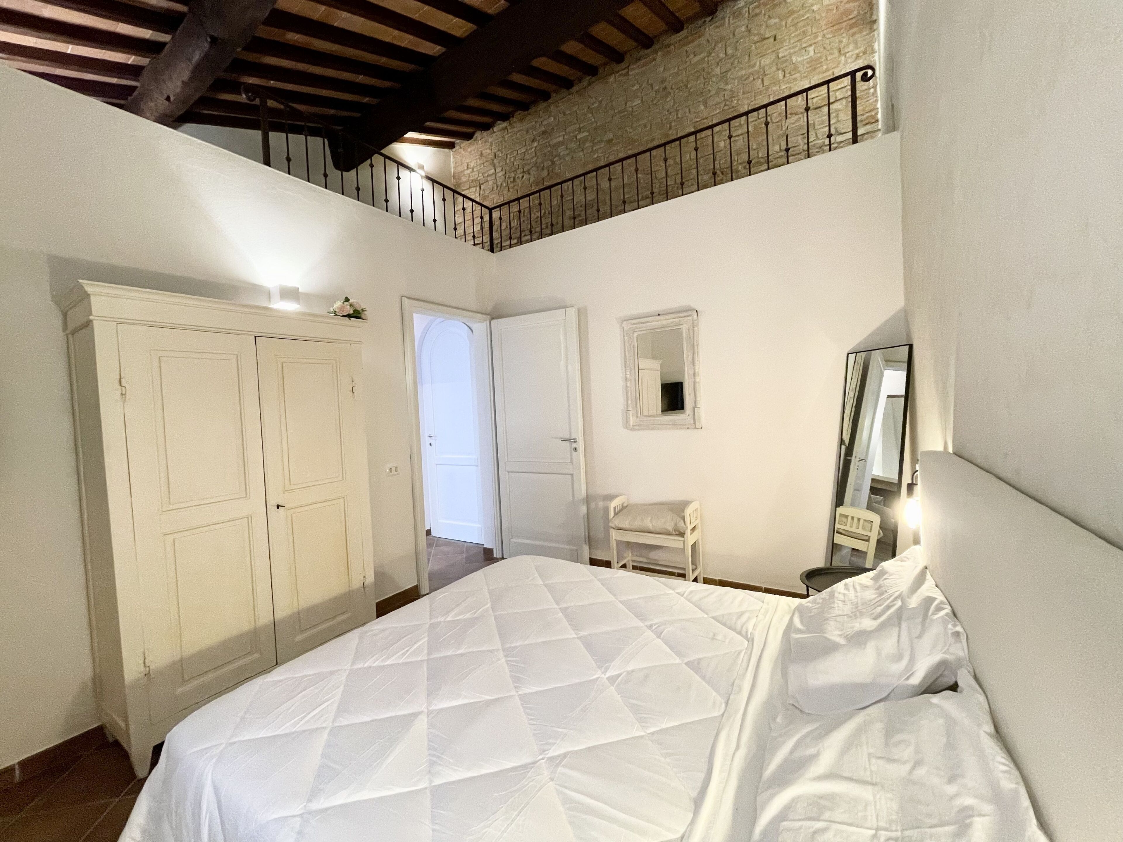 A bedroom with a large bed, a wardrobe, a small chair, a mirror, and a staircase leading to an upper level.