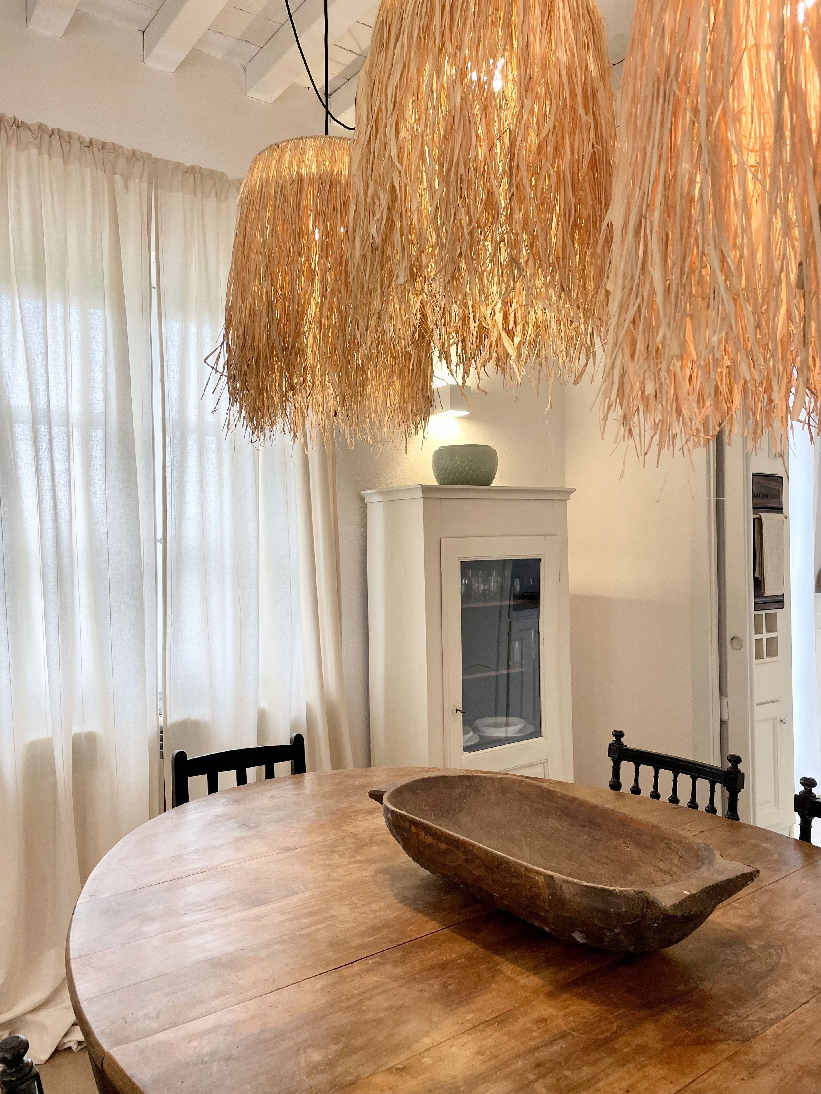 A dining area with a round wooden table, a woven hanging light fixture, and a white cabinet with glass doors.