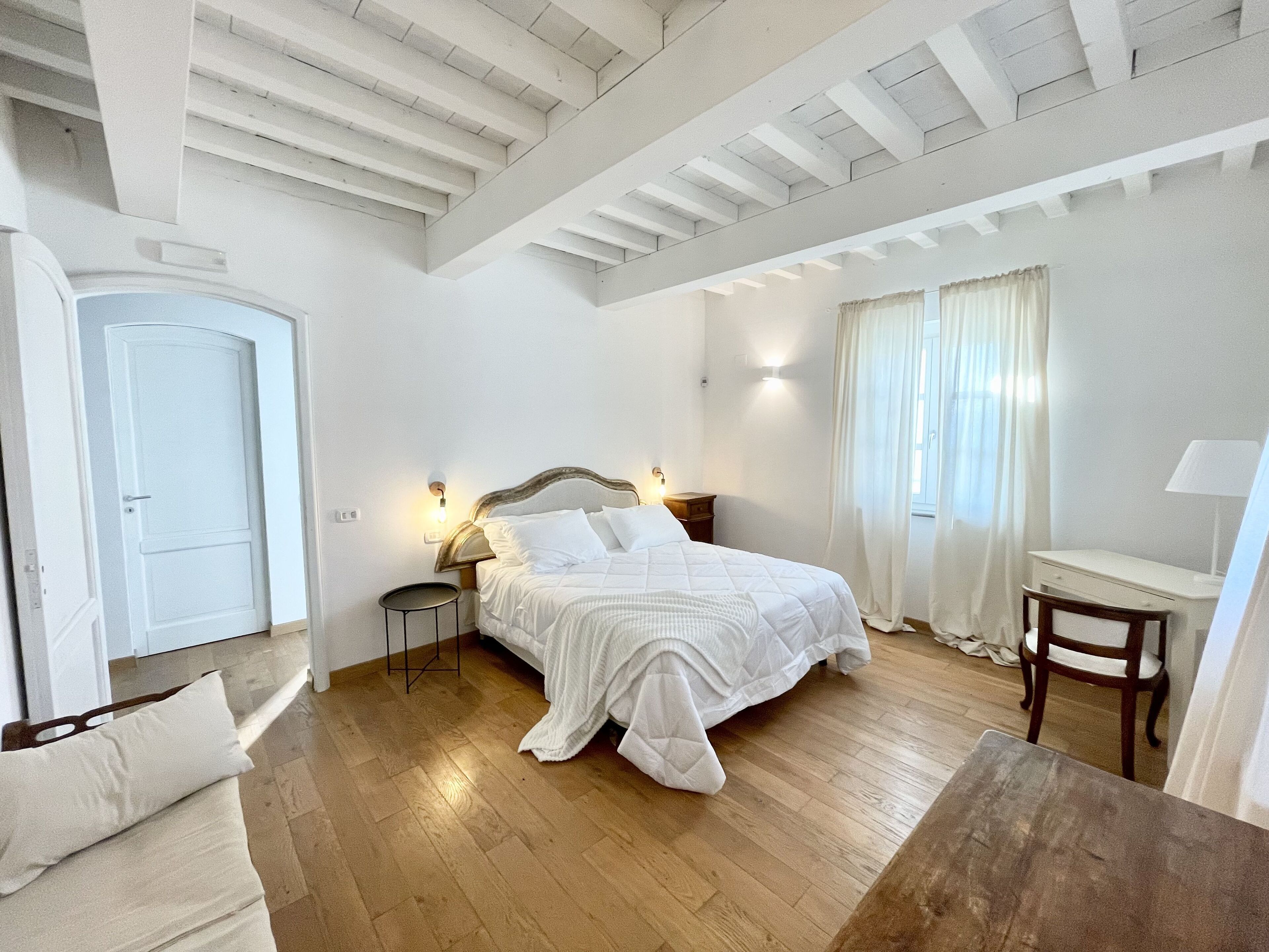 A bedroom with a large bed, wooden flooring, a desk, and a chair.