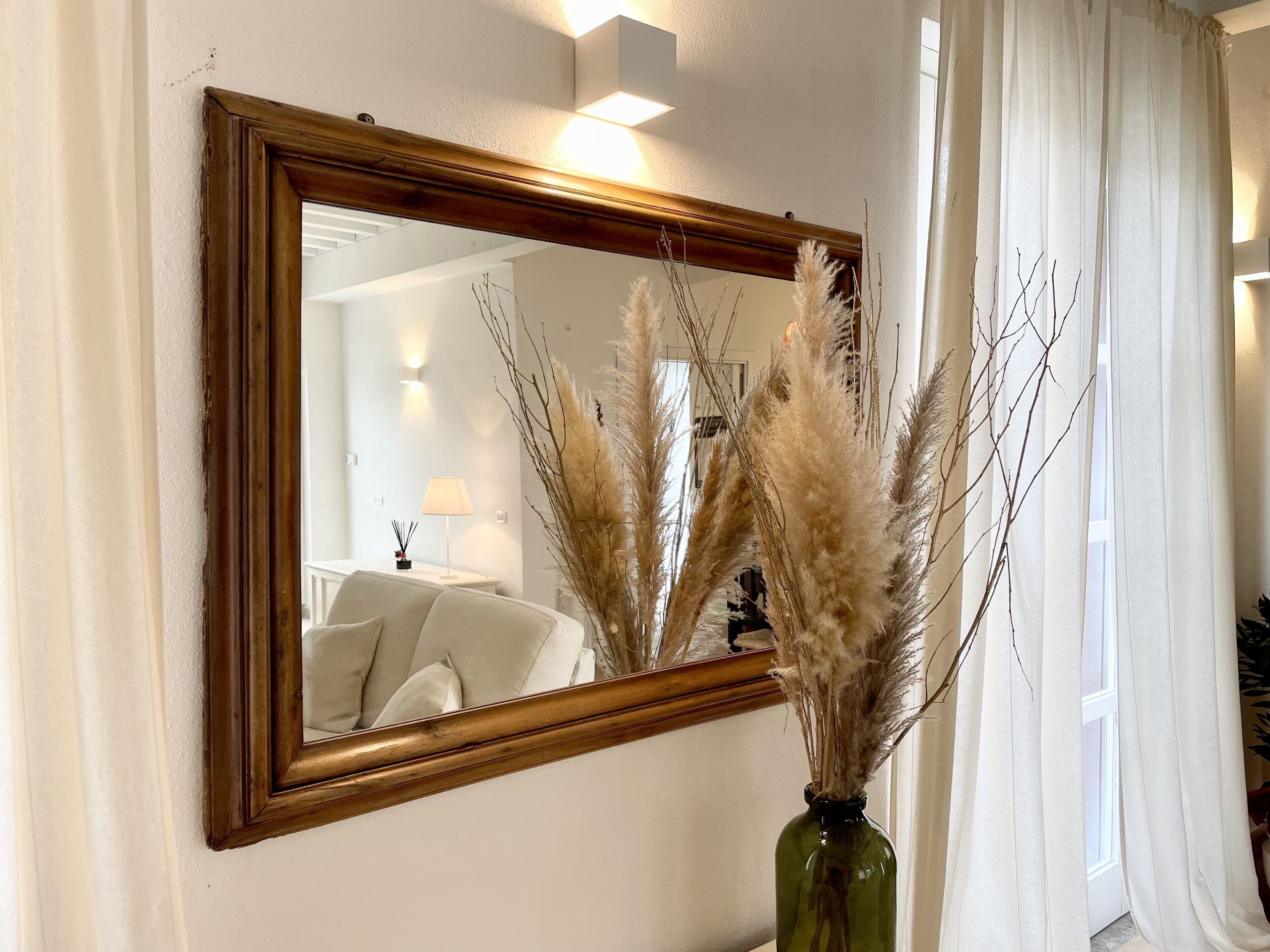 A mirror with a wooden frame on a wall, a vase with dried pampas grass in front of it, and a window with white curtains.