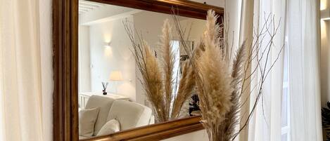 A mirror with a wooden frame on a wall, a vase with dried pampas grass in front of it, and a window with white curtains.