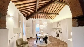 Un espace repas avec une table et des chaises, une cuisine avec des meubles blancs et un plafond voûté aux poutres apparentes.