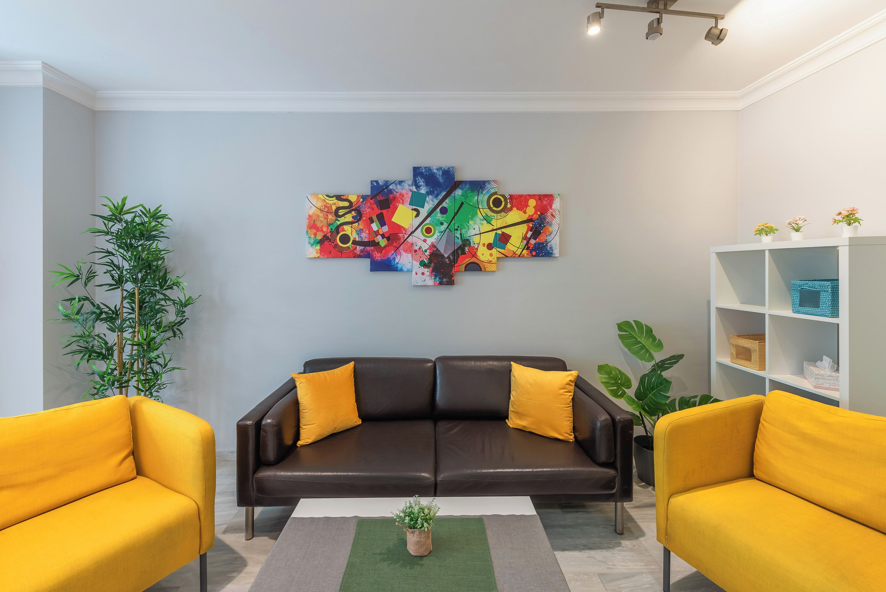 A modern living room with a black leather sofa, yellow armchairs, a colorful abstract painting, and a white shelving unit.