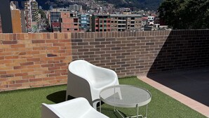 Terraço no telhado com cadeiras brancas e uma mesa de metal, com vista para a paisagem urbana e montanhas.