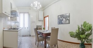 Une cuisine moderne avec une cuisinière à gaz, des armoires blanches et un lustre. On y trouve un coin repas avec une table et des chaises en bois, ainsi qu’une plante en pot.