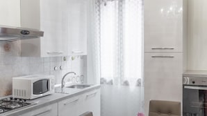 Une cuisine moderne avec des armoires blanches, un four à micro-ondes, un évier et une cuisinière. On y trouve un coin repas avec une table et des chaises, ainsi qu’une plante en pot sur la table.