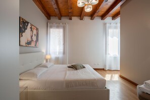 Un dormitorio con una cama grande, vigas de madera, un cuadro en la pared y dos ventanas con cortinas.