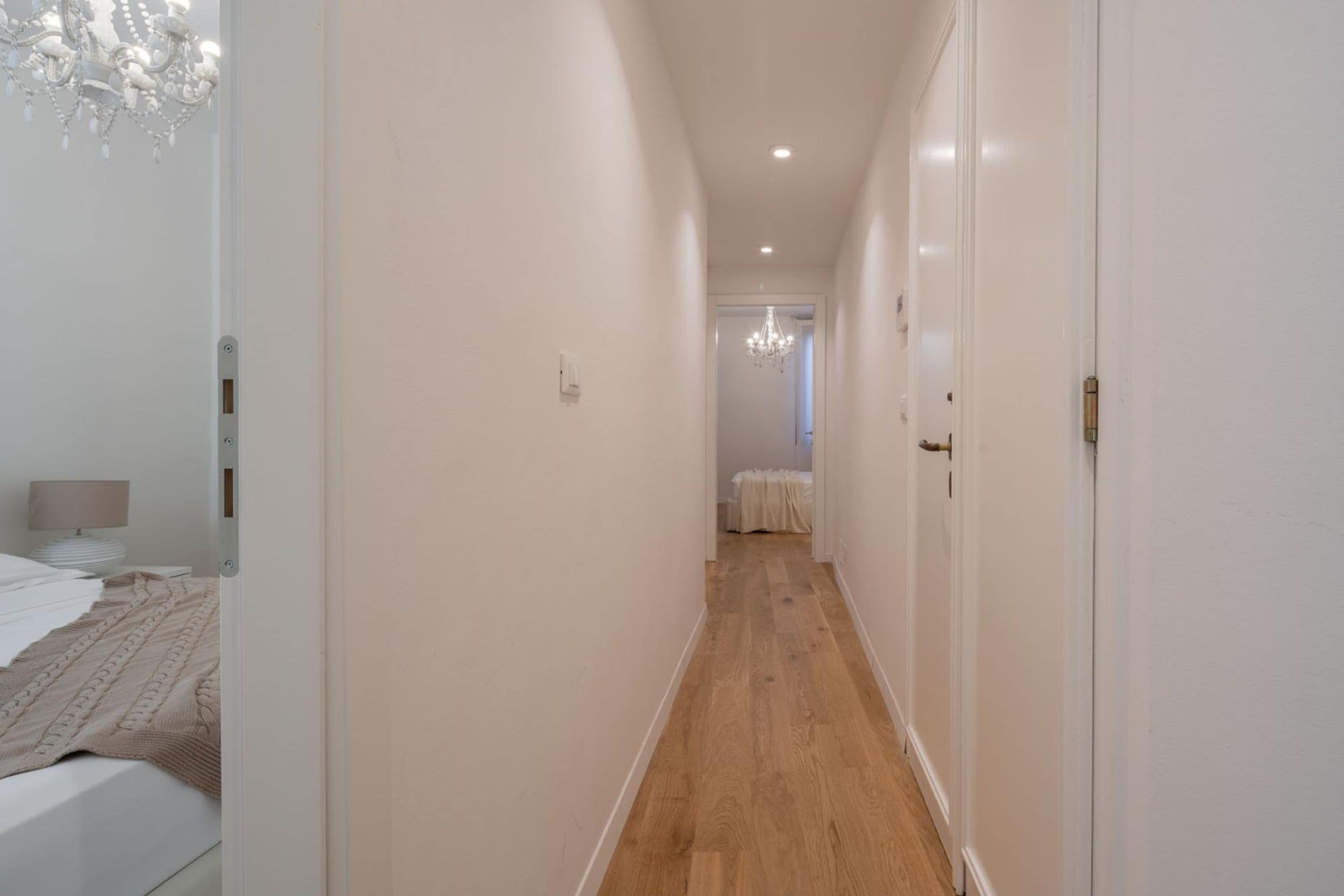A narrow hallway with wooden flooring and white walls, leading to a room with a bed and a chandelier.