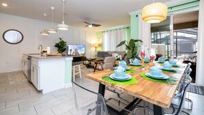 A modern kitchen with a central island, dining area with a wooden table set for six, and a large window with curtains. - Vibrant Themed Home Prime Location (Kissimmee)