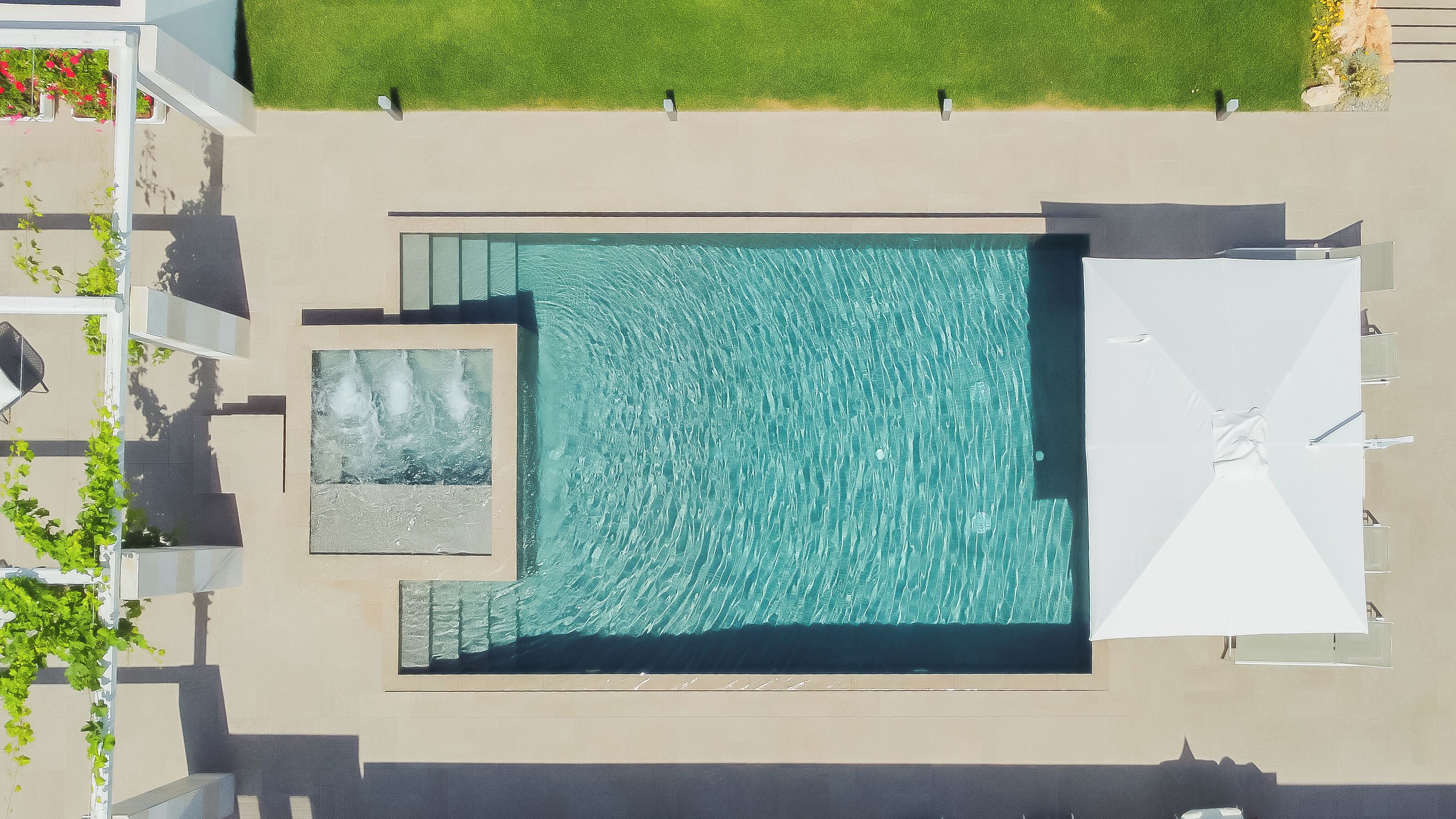 An aerial view of a rectangular swimming pool with a small adjacent water feature, surrounded by a paved deck and a grassy area.