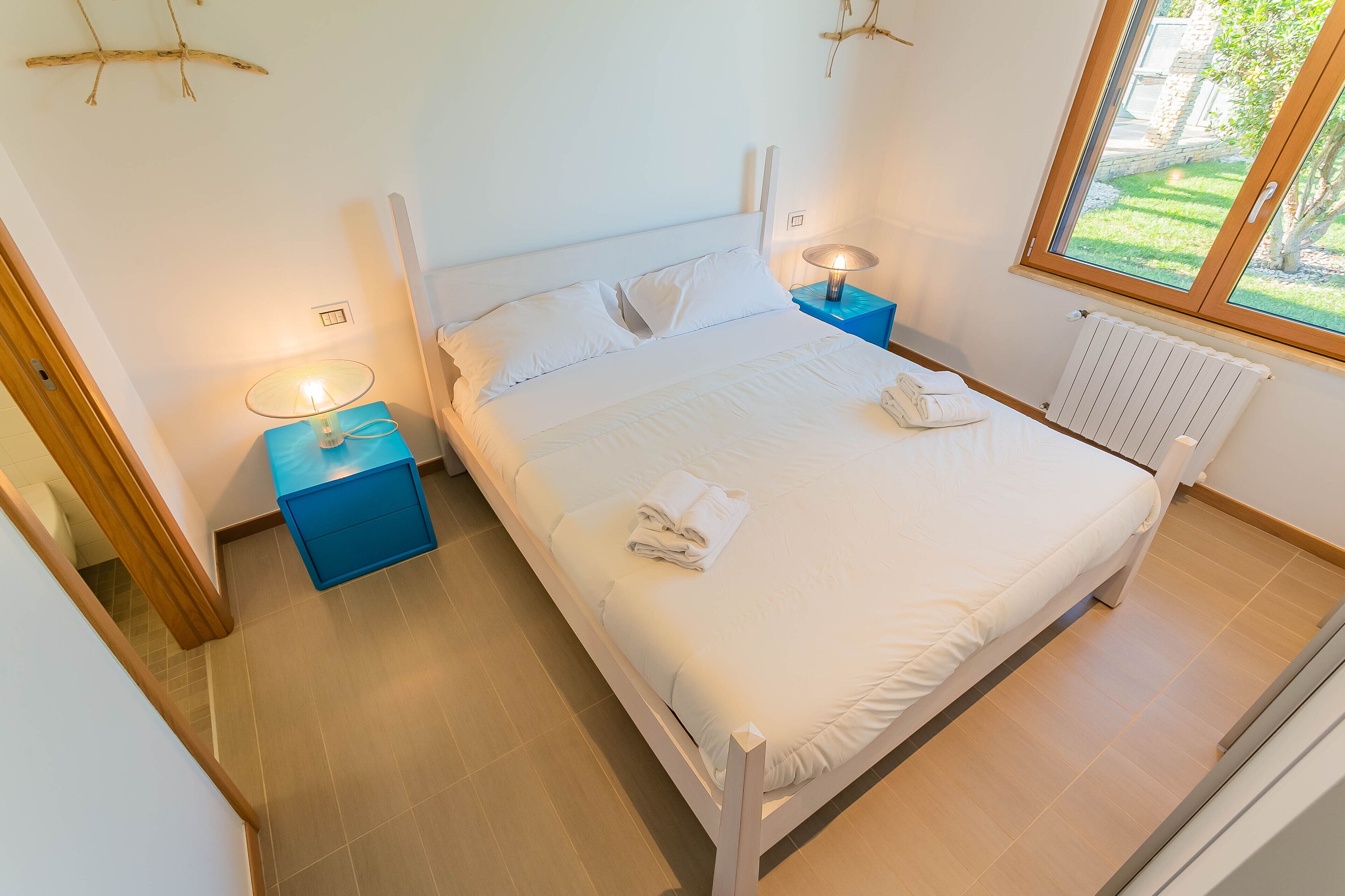 A neatly made bed with white linens and towels, a bedside lamp, and a view of a garden through a window.