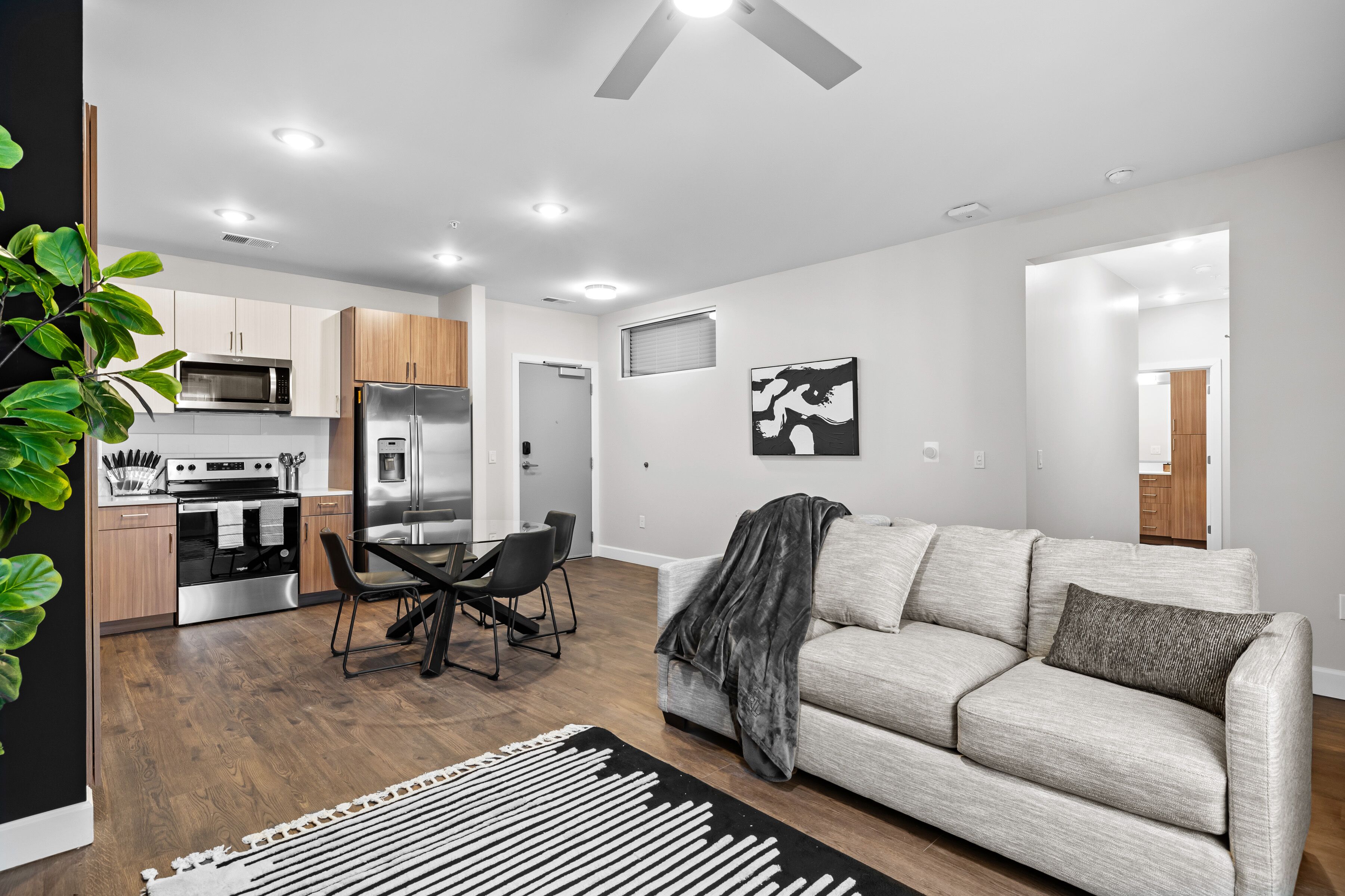 A modern living room with a sofa, a dining table with chairs, a kitchen area with appliances, and a large plant.