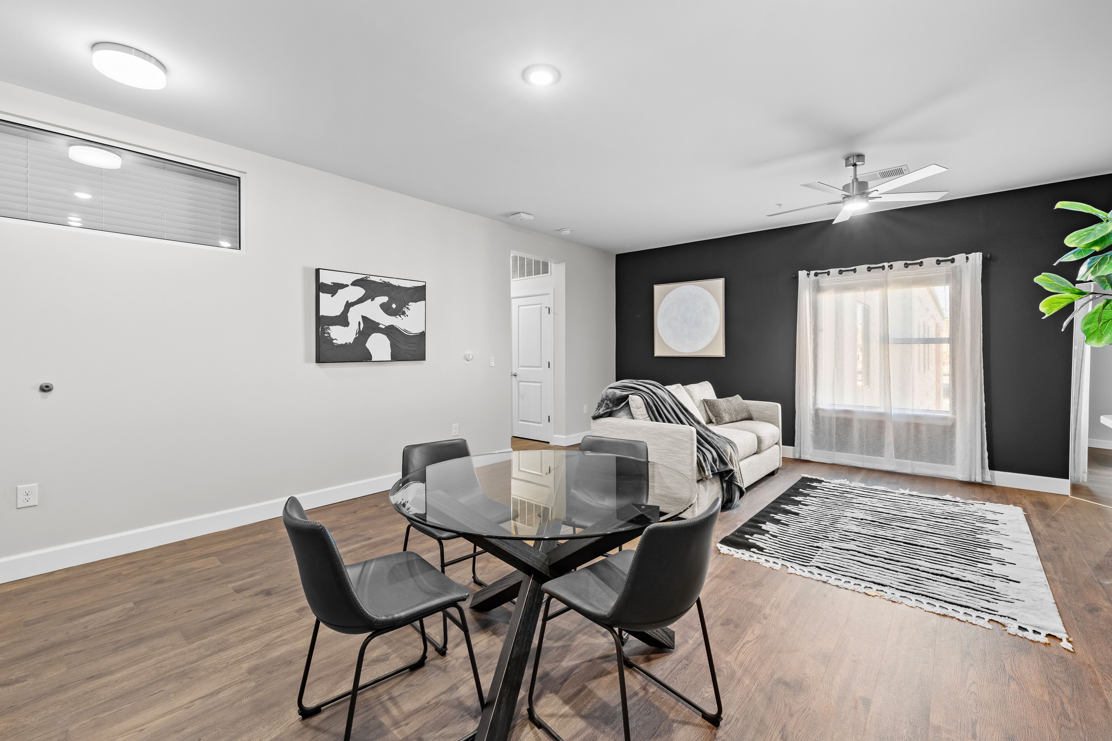 A modern living room with a glass dining table, black chairs, a sofa, and a large window with curtains.