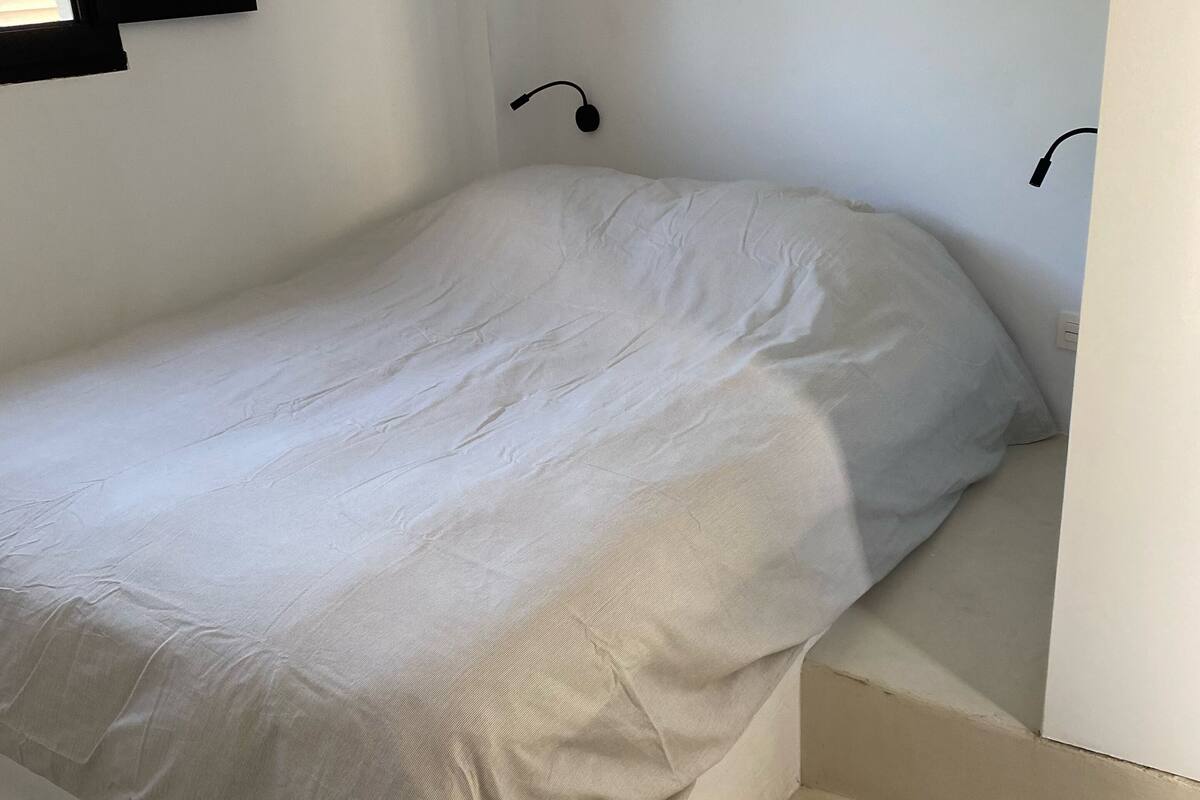 A small, single-bed room with a plain white bedspread, a black wall-mounted lamp, and a white wall.