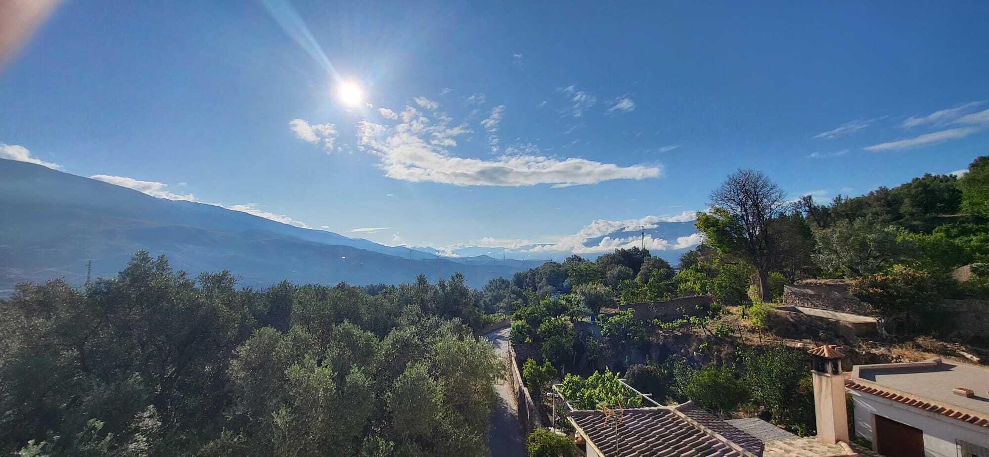 A mountainous landscape with a clear sky and a sun shining. There are trees and a building with a tiled roof.