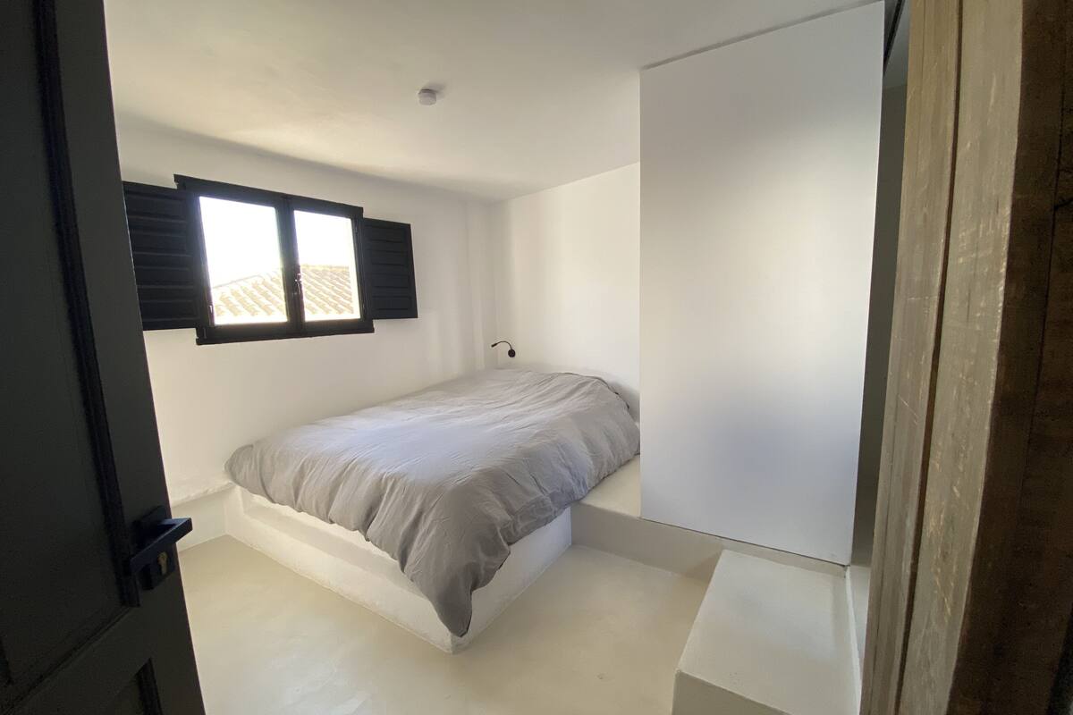 A small, single-bed room with a window, white walls, and a simple, uncluttered layout.