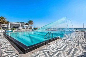 A swimming pool with clear blue water, surrounded by a tiled deck and a covered area with lounge chairs.