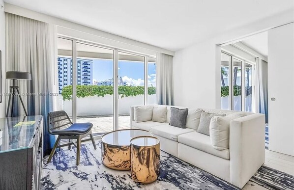 A modern living room with a white sofa, two round coffee tables, a glass desk, and a chair. Large windows offer a view of buildings and greenery.