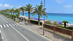 Una carretera costanera amb palmeres, pas per als vianants i una platja amb ombrelles blaves.