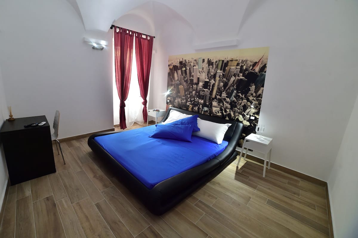 A bedroom with a bed, a nightstand, a wall-mounted air conditioner, a red curtain, and a cityscape mural on the wall.