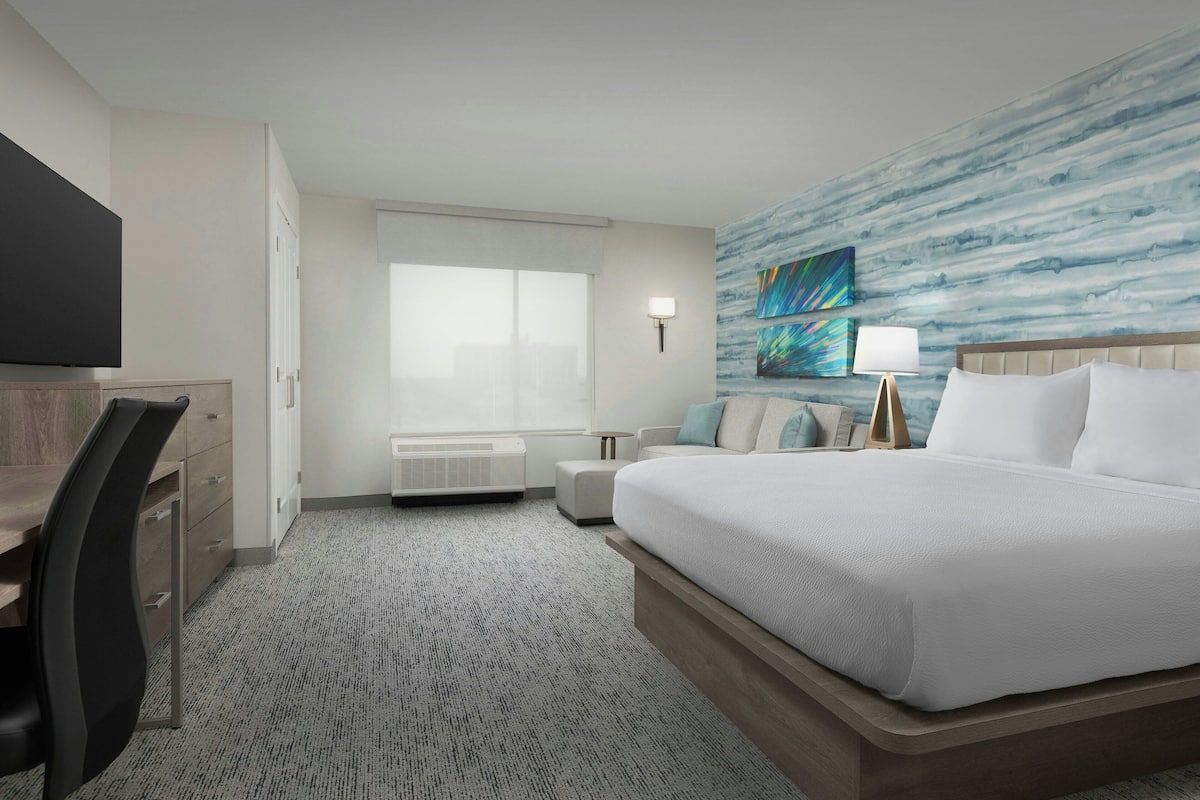 A hotel room with a large bed, a desk, a chair, a TV, a window, an air conditioning unit, and a wall with a painting.
