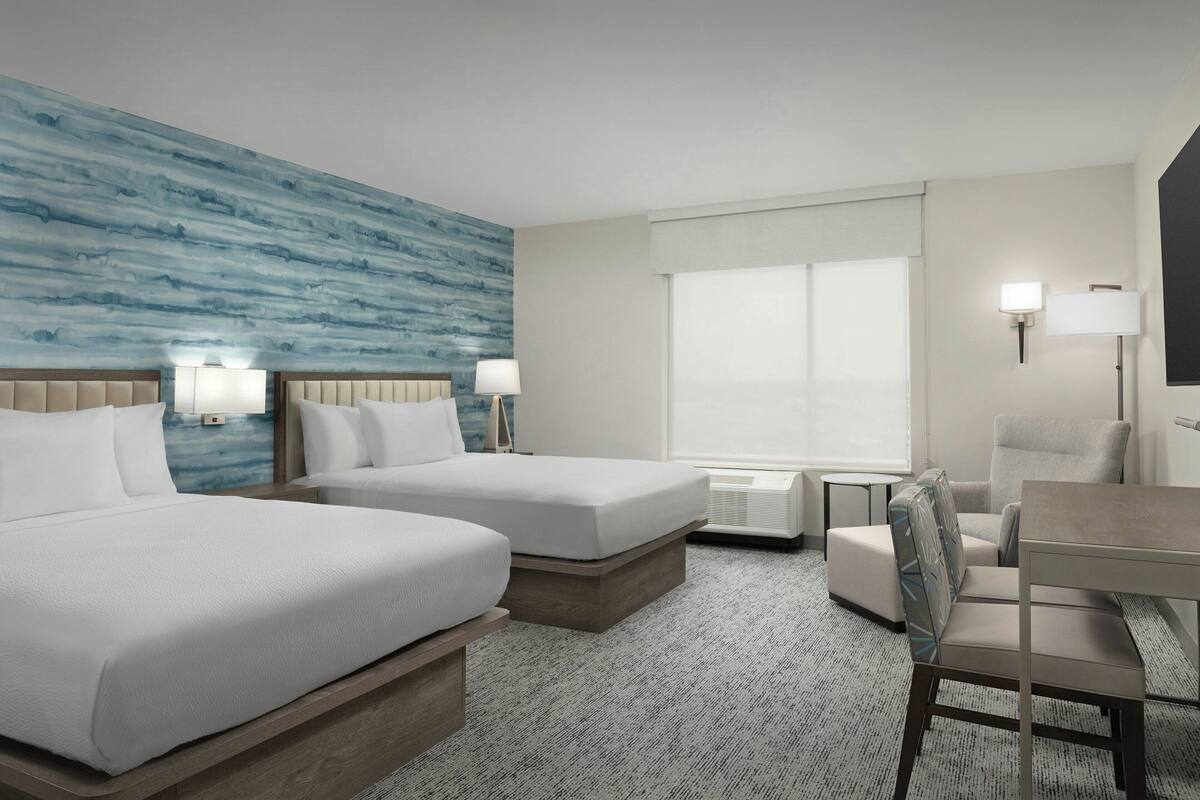 A hotel room with two beds, a desk, a chair, a large window, and a wall with a blue pattern.