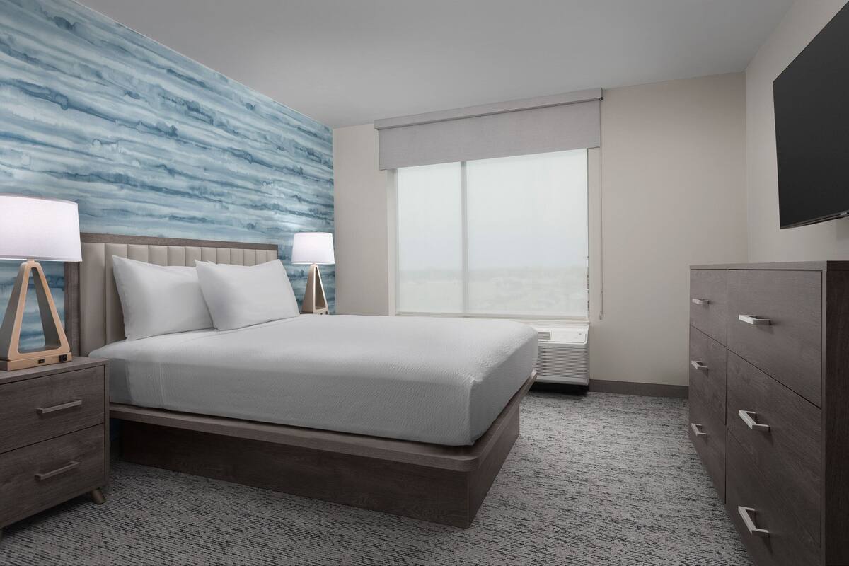 A modern hotel room with a large bed, a nightstand, a dresser, and a television on the wall.