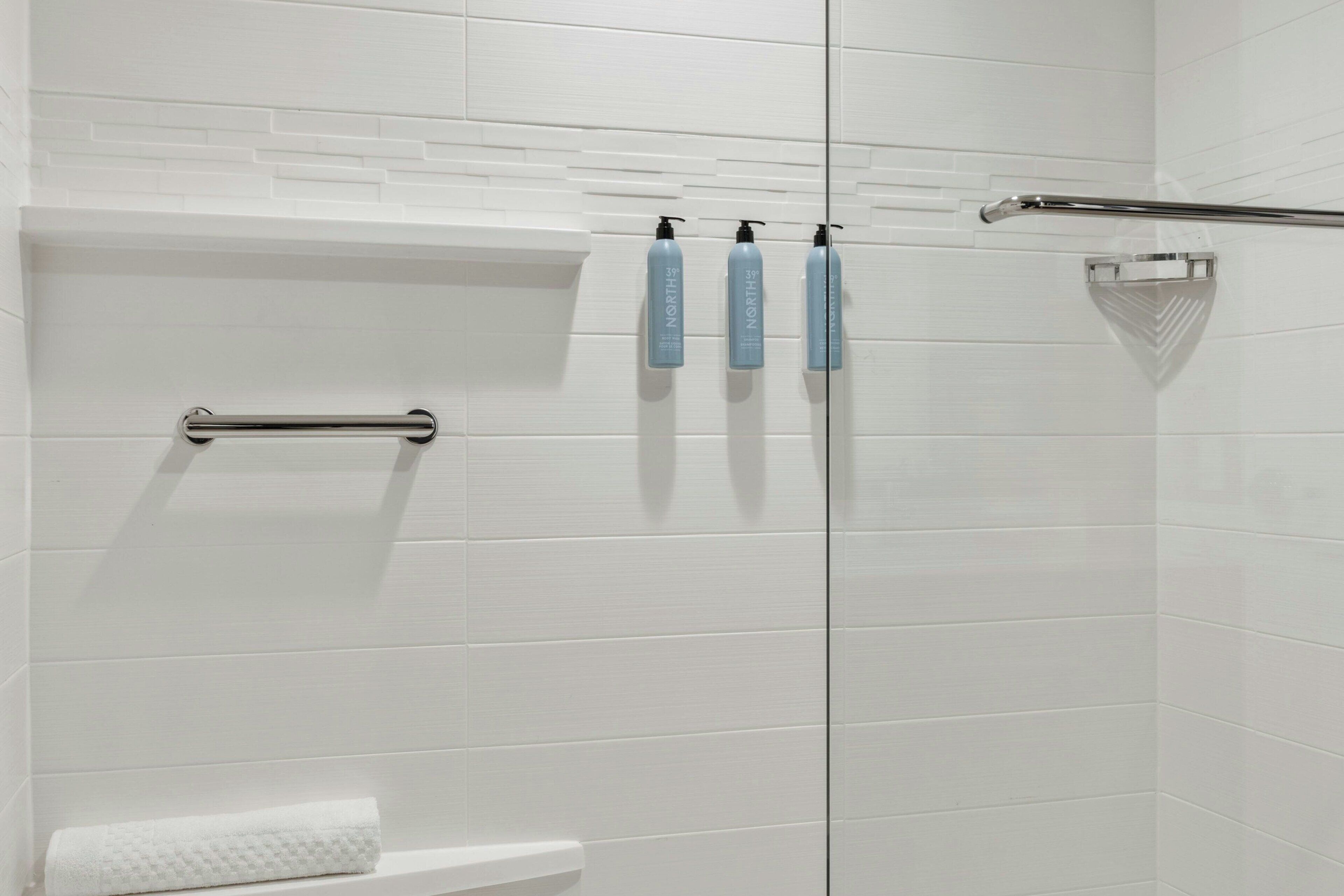 A modern bathroom with white subway tiles, a glass shower enclosure, a handrail, and three bottles of shampoo on a towel rack.