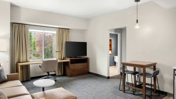 Ein Hotelzimmer mit einer Couch, einem kleinen runden Tisch, einem Fernseher, einem Schreibtisch mit Stuhl sowie einer Bar mit Hockern.
