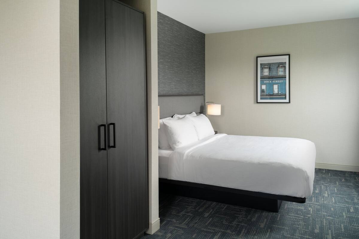 Une chambre d’hôtel avec un lit, une armoire, une lampe fixée au mur et un tableau encadré accroché au mur.