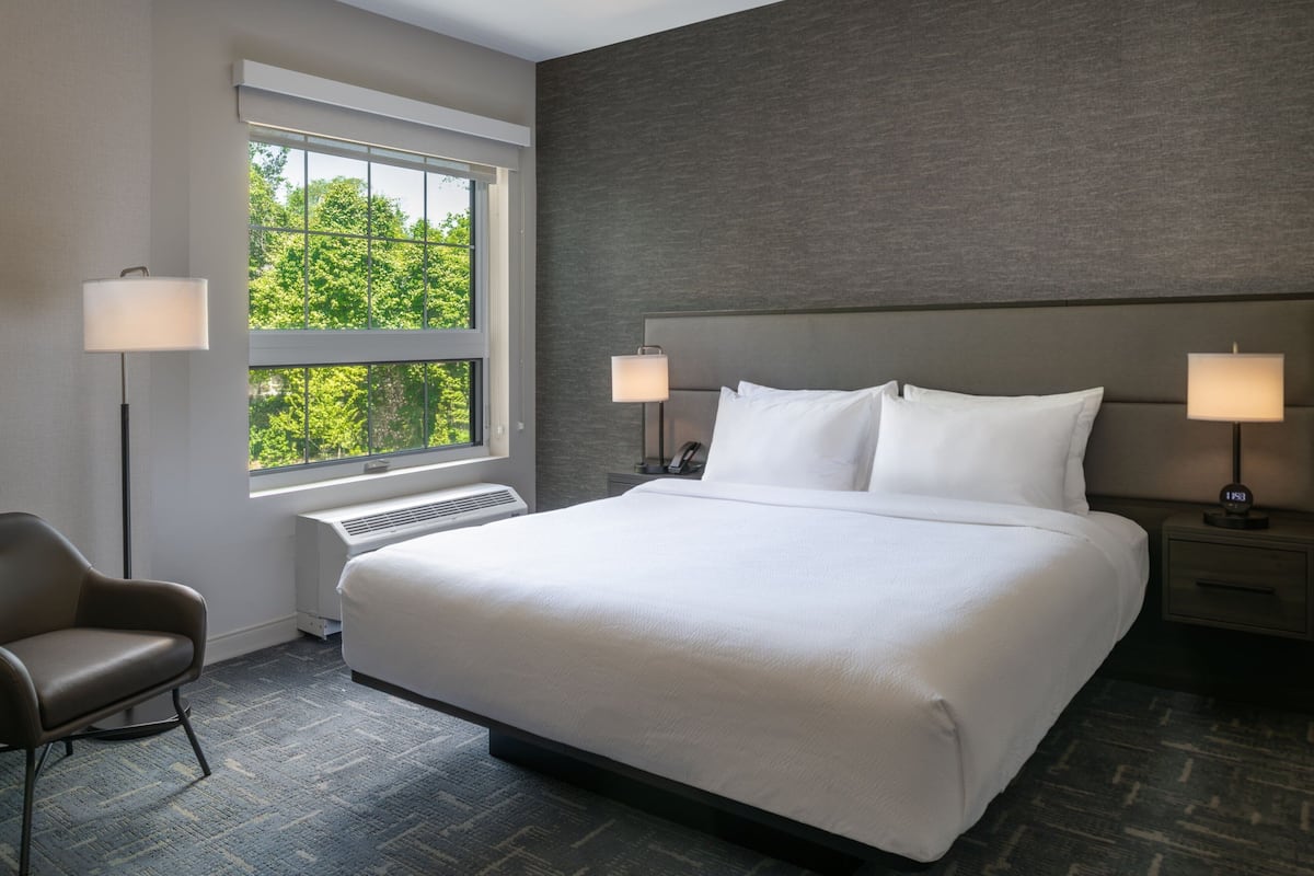 Une chambre d’hôtel moderne avec un grand lit, deux lampes de chevet, une chaise et une fenêtre donnant sur des arbres.