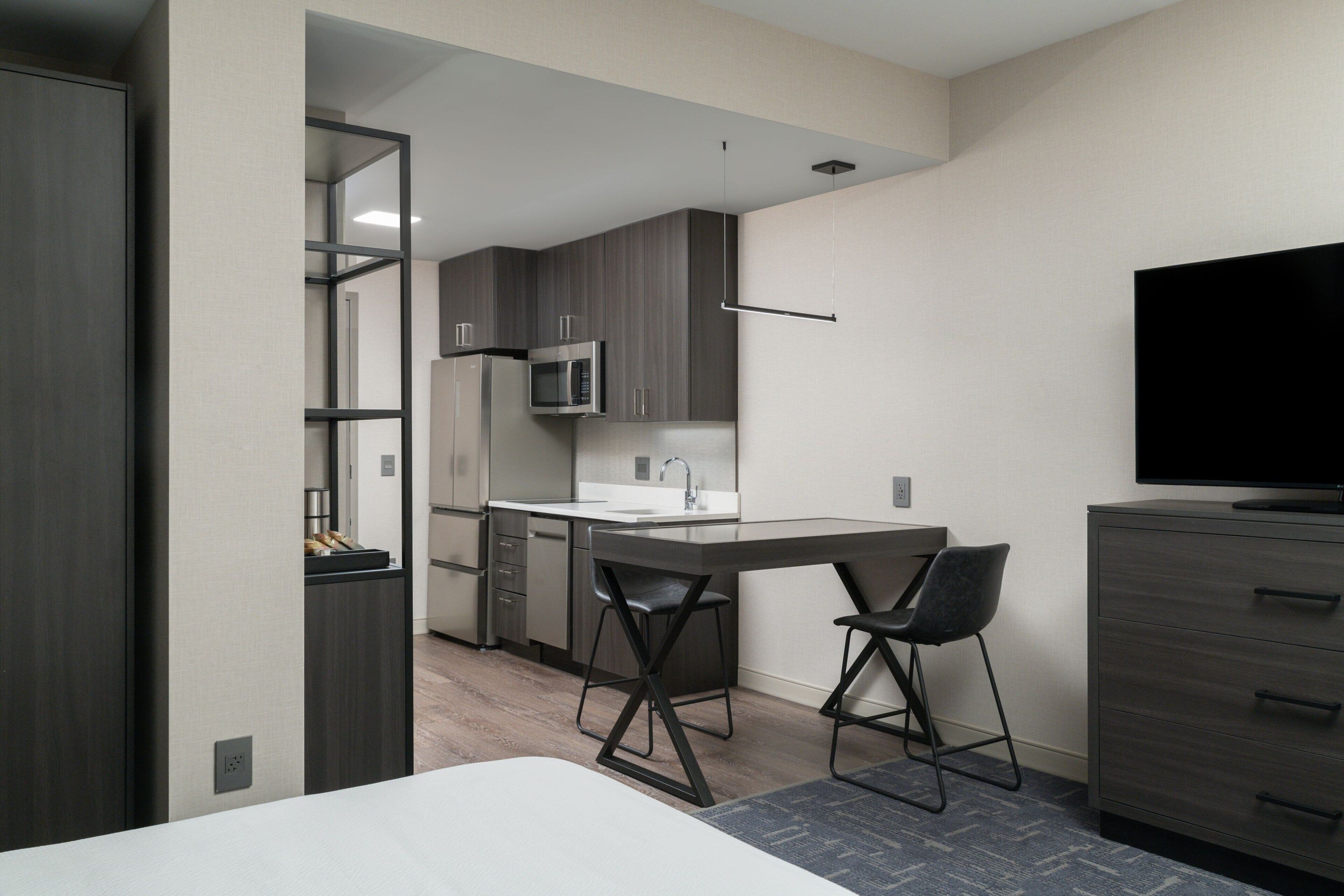 A modern hotel room with a bed, a desk with two chairs, a flat-screen TV, a kitchenette with a microwave, and a wardrobe.