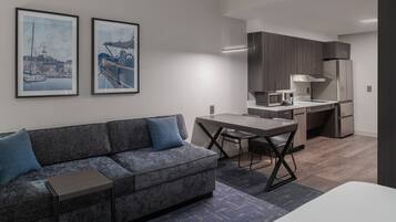A modern hotel room with a grey sofa, a small table, a dining area with a chair, a kitchen area with a microwave, and a refrigerator.