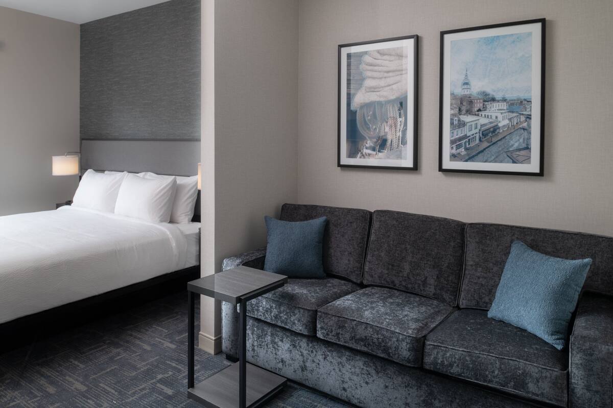 A modern hotel room with a bed, a grey sofa, two framed artworks, and a small table.