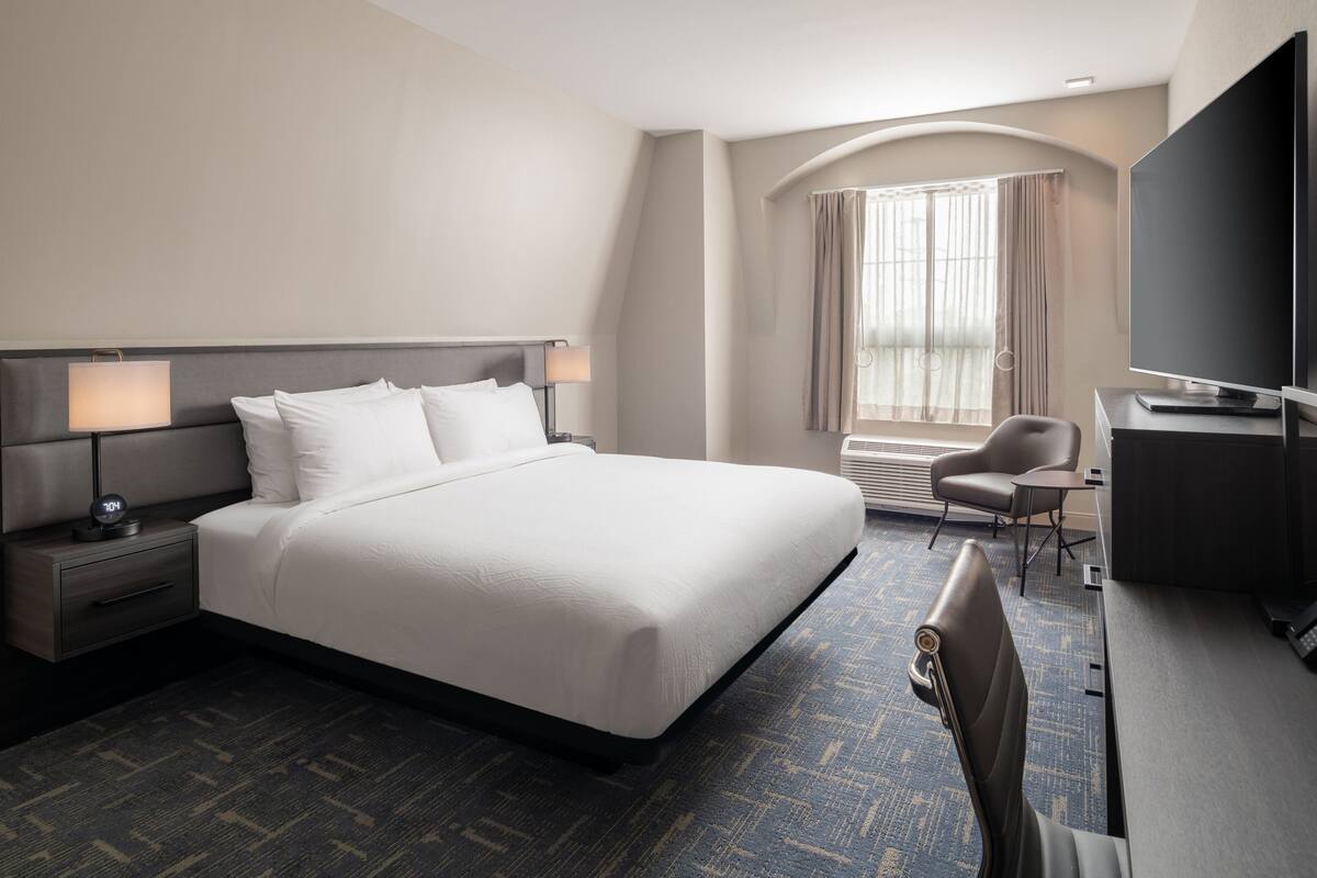 A hotel room with a large bed, a desk, a chair, a TV, and a window with curtains.
