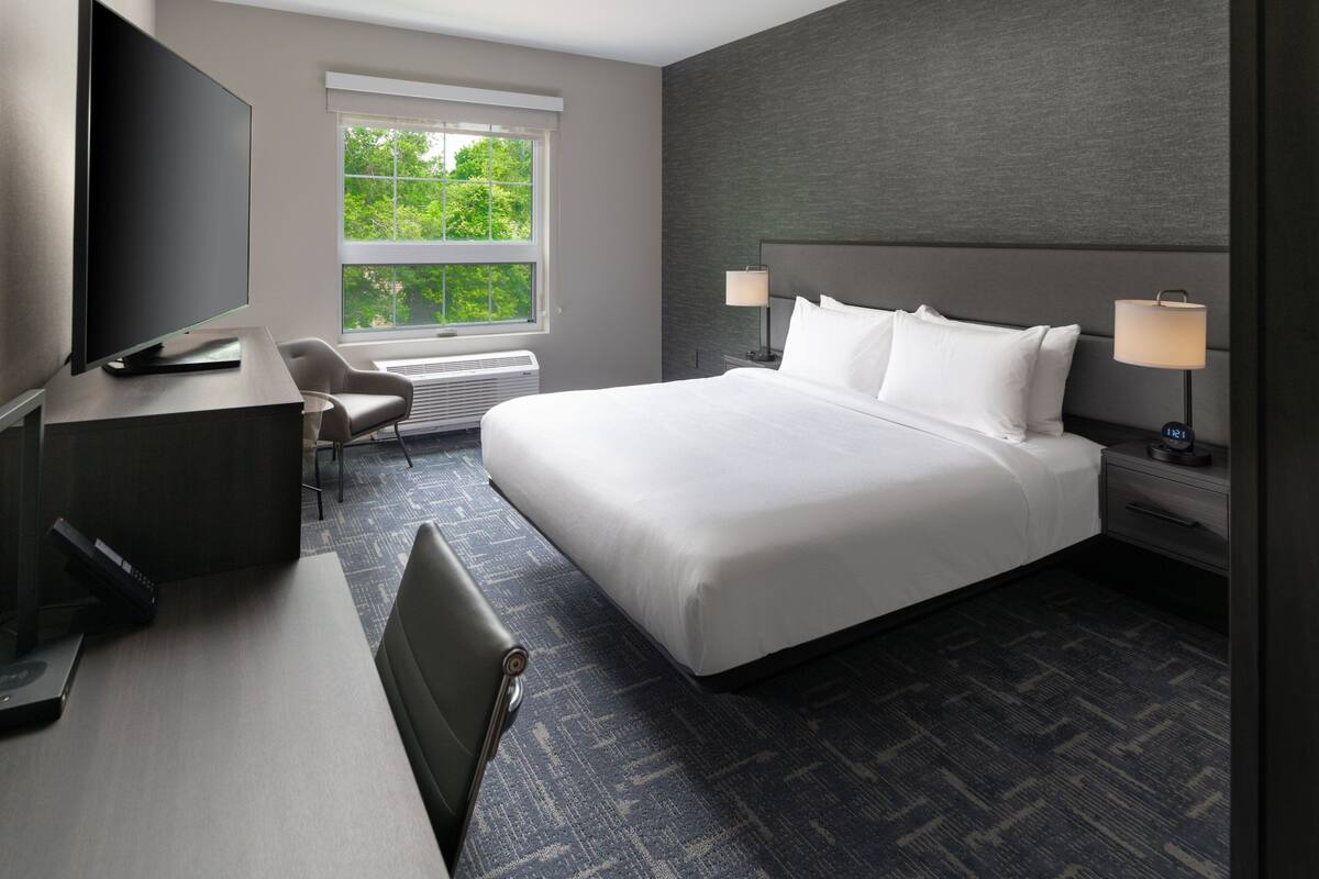 A modern hotel room with a large bed, a desk, a chair, a TV, and a window with a view of trees.
