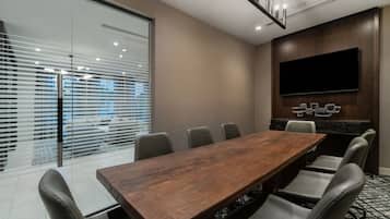 A conference room with a long wooden table, six chairs, a flat-screen TV mounted on the wall, and a glass partition with blinds.